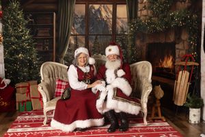Santa and Mrs. Claus sit together in a festive holiday setting with Christmas trees, gifts, and a cozy fireplace backdrop.