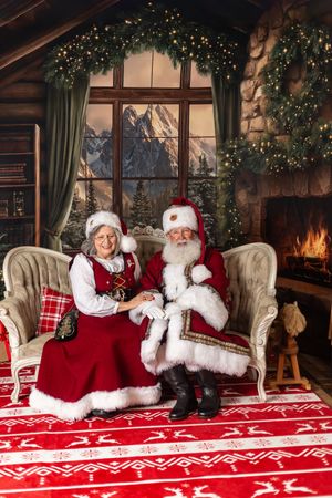 Santa and Mrs. Claus sitting together on a elegant sofa in a cozy Christmas decorated cabin with fireplace.