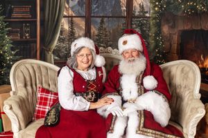 A cheerful couple dressed as Santa Claus and Mrs. Claus sitting together on an elegant chair in a festive Christmas setting.