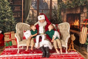 Two children sit on Santa Claus's lap by a cozy fireplace surrounded by Christmas trees and gifts.