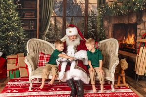 Two young boys sit on a couch with Santa Claus by a fireplace and Christmas tree in a festive holiday setting.