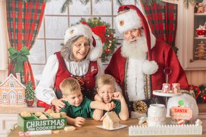Santa and Mrs. Claus smile with two children decorating Christmas cookies and a gingerbread house.