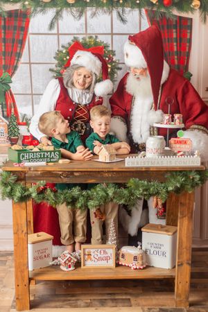 Two children bake Christmas cookies with Santa and Mrs. Claus at a festive wooden table decorated with garland and holiday signs.