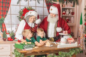 Santa and Mrs. Claus make Christmas cookies with two young children at a holiday decorated kitchen counter.