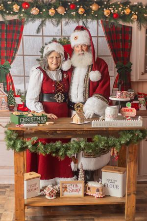 Santa and Mrs. Claus smile together at a festive baking table decorated with Christmas cookies and holiday decor.