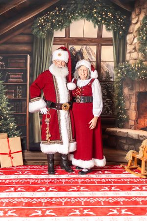 Santa and Mrs. Claus in red velvet suits pose together in a festive Christmas workshop setting with decorated tree.