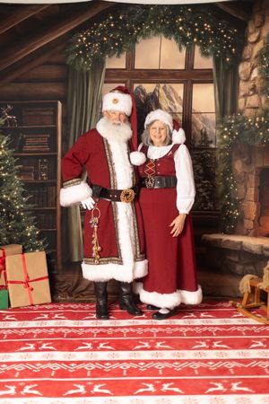 Santa and Mrs. Claus in dark red velvet outfits stand together in a cozy holiday-decorated Christmas workshop scene.