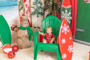 Toddler in red Hawaiian shirt sits in green Adirondack chair with Christmas surfboards in tropical holiday beach scene.