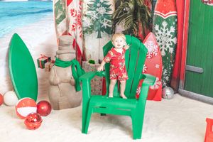 Toddler in red Christmas outfit standing on a green beach chair in a tropical holiday photo setting.