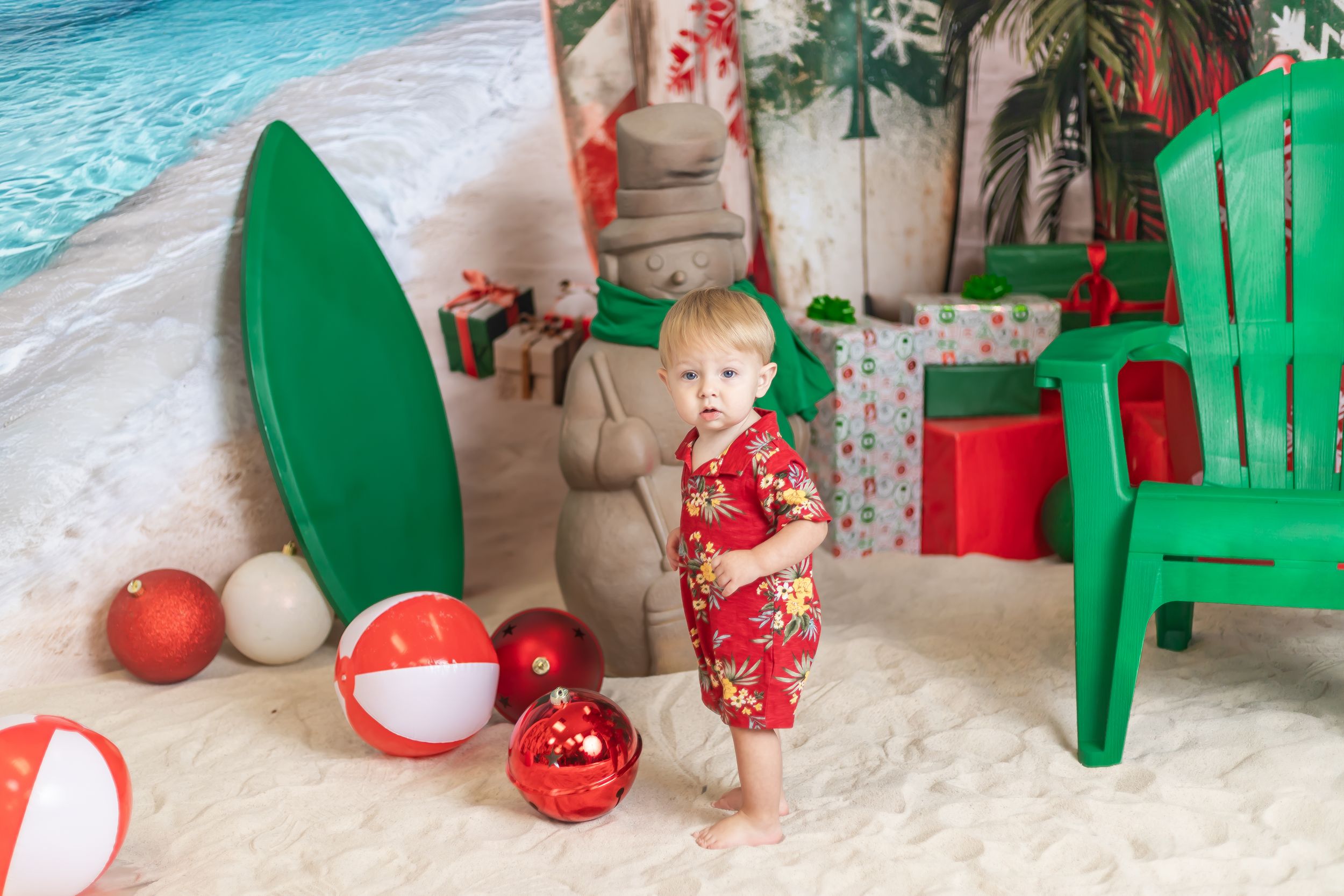 Toddler in red floral outfit stands on sandy beach Christmas backdrop with colorful ornaments, surfboard, and holiday gifts.