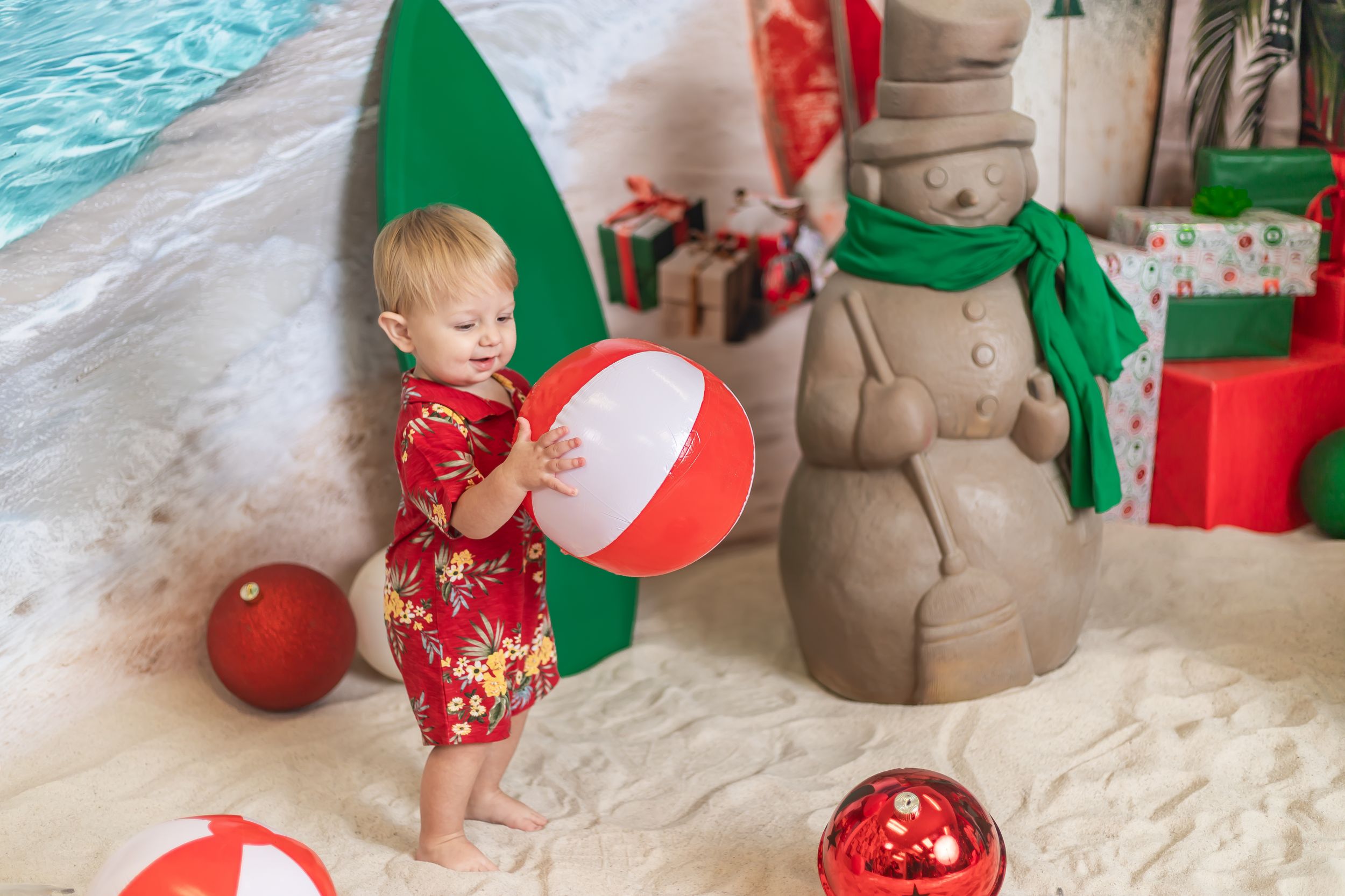 Toddler in red floral outfit plays with beach ball near sand snowman and Christmas gifts in festive beach holiday scene.