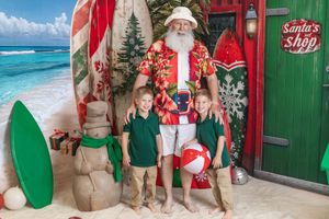 Two children pose with a beach-themed Santa Claus in front of surfboards and Christmas decorations at Santa's Shop.