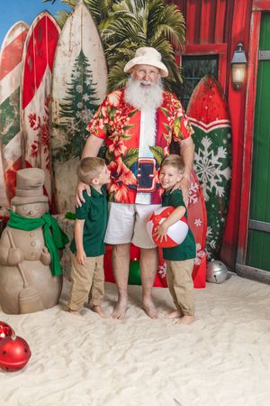 Santa in Hawaiian shirt and hat poses with two children on a sandy beach Christmas set with surfboards.
