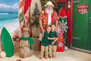 Two boys pose with a beach Santa Claus near surfboards and a Santa's Shop sign in a tropical Christmas scene.