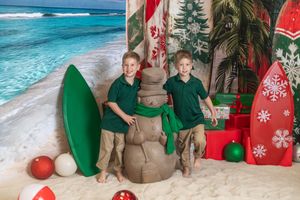 Two boys in green polo shirts pose with a sand snowman on a beach Christmas backdrop with surfboards.