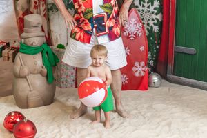 Toddler holding beach ball in Christmas-themed tropical photo shoot with snowman decor and holiday ornaments.