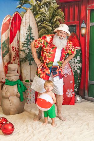 Santa Claus in Hawaiian shirt and white shorts poses with toddler on sandy beach Christmas theme backdrop with surfboards.