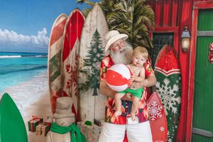 Santa Claus in Hawaiian shirt holds toddler with beach ball in tropical Christmas photo backdrop.