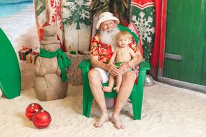 Santa Claus in Hawaiian shirt holds baby on beach-themed Christmas set with surfboard and snowman decorations.