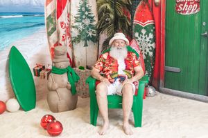 Santa Claus sitting in a green chair on a tropical beach-themed Christmas set with surfboards and palm trees.