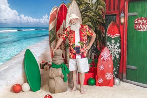 Santa Claus in Hawaiian shirt and shorts poses on tropical beach with surfboards and Christmas decorations.