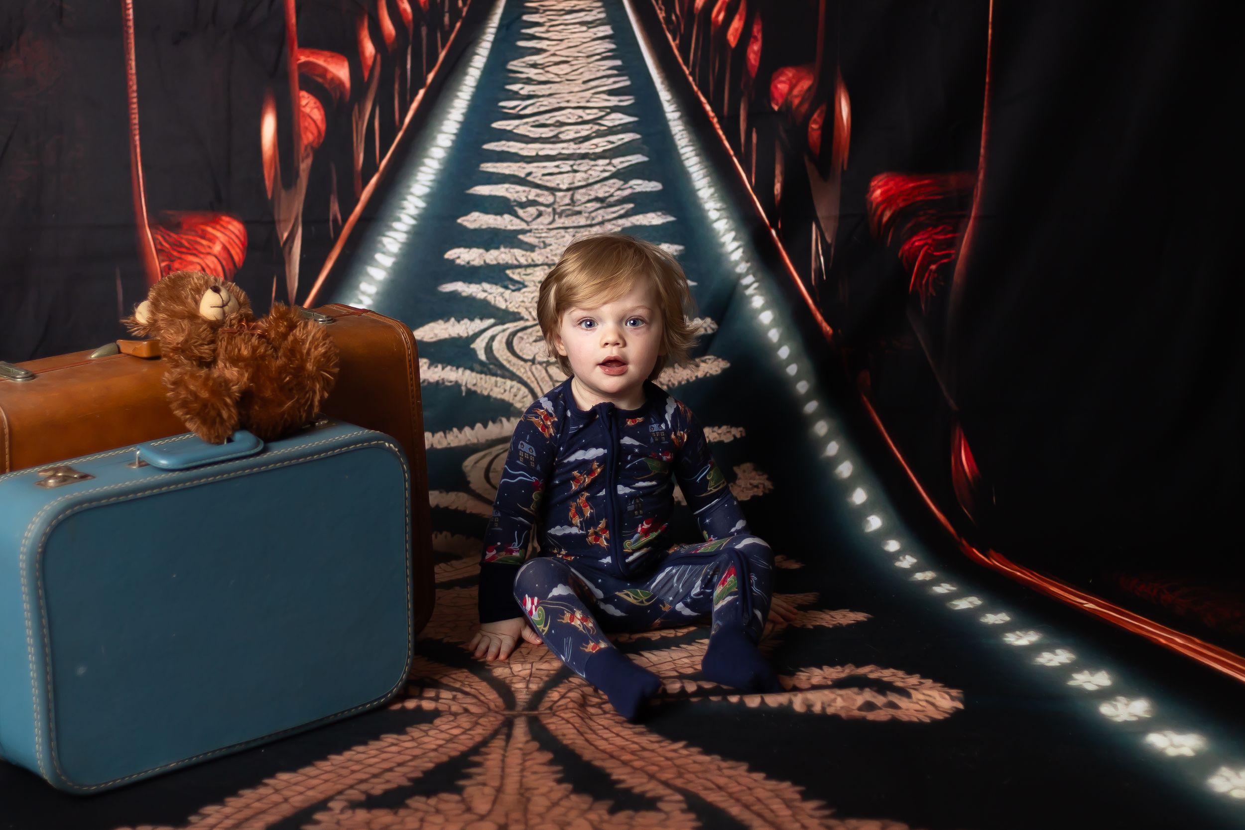 Toddler in navy pajamas sits on ornate carpet between vintage suitcases in a dramatic dark tunnel setting.