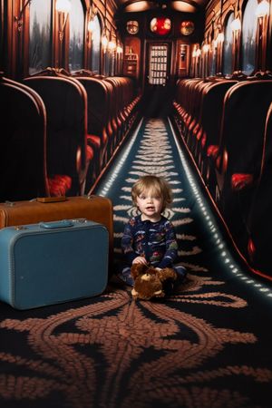 A toddler sits on a patterned carpet in a vintage train aisle with red seats, luggage, and a teddy bear nearby.