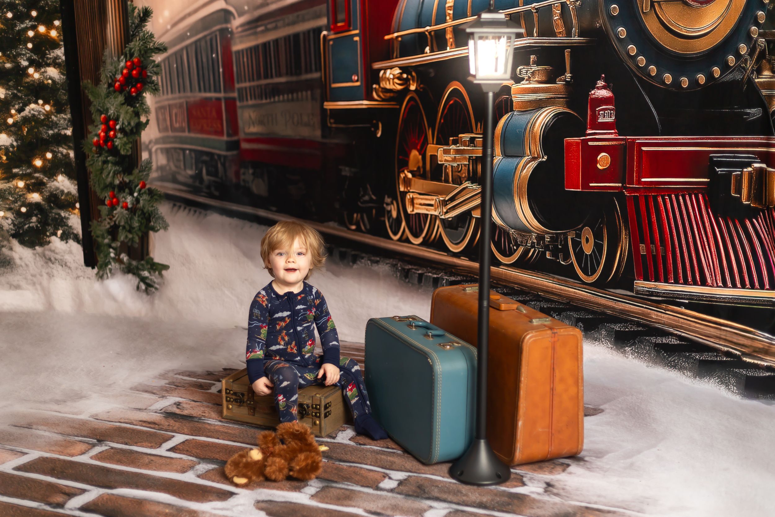 Young toddler in festive pajamas sits beside vintage suitcases near a Christmas train backdrop with a glowing lamp post.