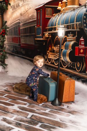 Toddler in holiday pajamas poses with vintage suitcases in front of a Polar Express train backdrop.