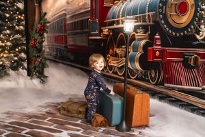 A young child in festive pajamas poses with vintage luggage beside a Christmas train station backdrop with snow.