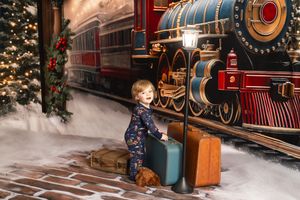 Young child in festive pajamas sitting on vintage luggage beside a Christmas train station backdrop with holiday decorations.