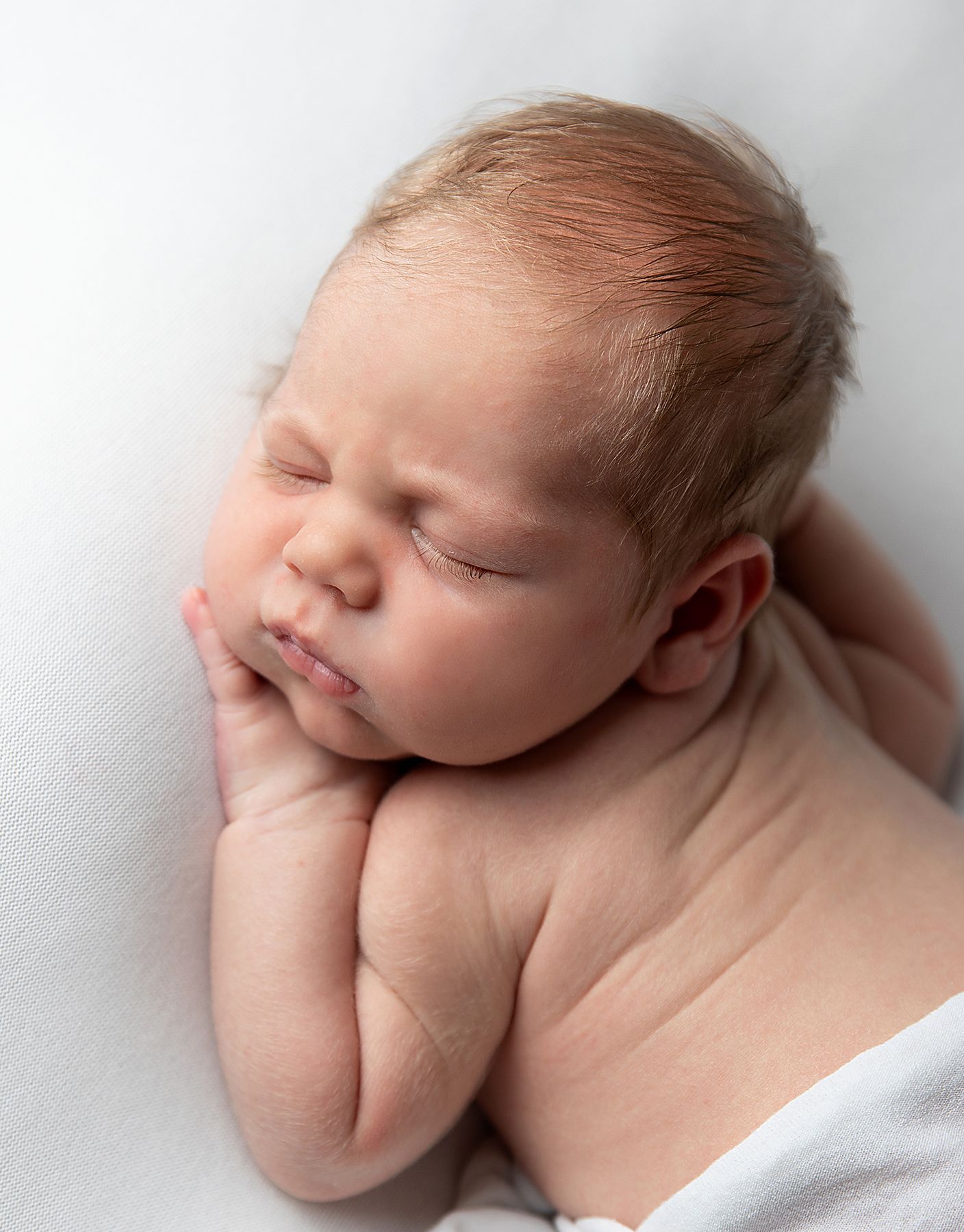 Sleeping newborn infant rests peacefully against white background with hands tucked under chin. During a newborn photoshoot in Bexley
