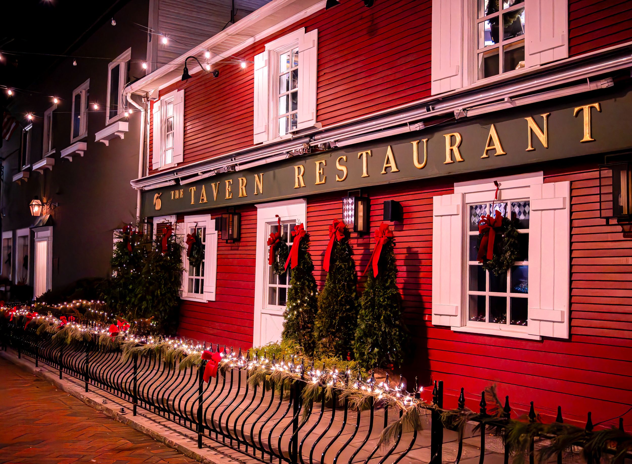 Charming red tavern restaurant with white trim windows and twinkling string lights decorating the entrance at night.