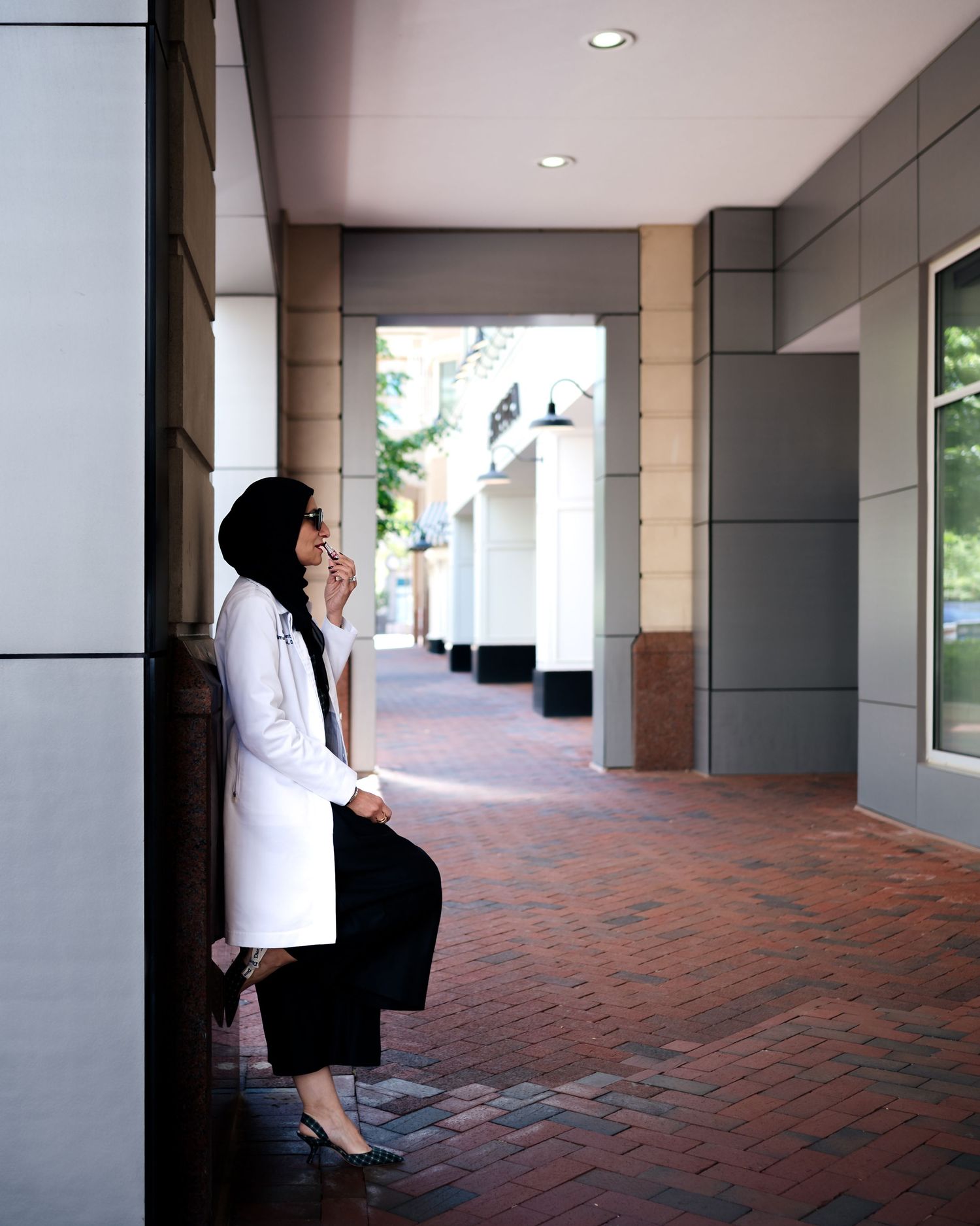 A woman in a white coat and black hijab leans against a modern building wall on a brick walkway.