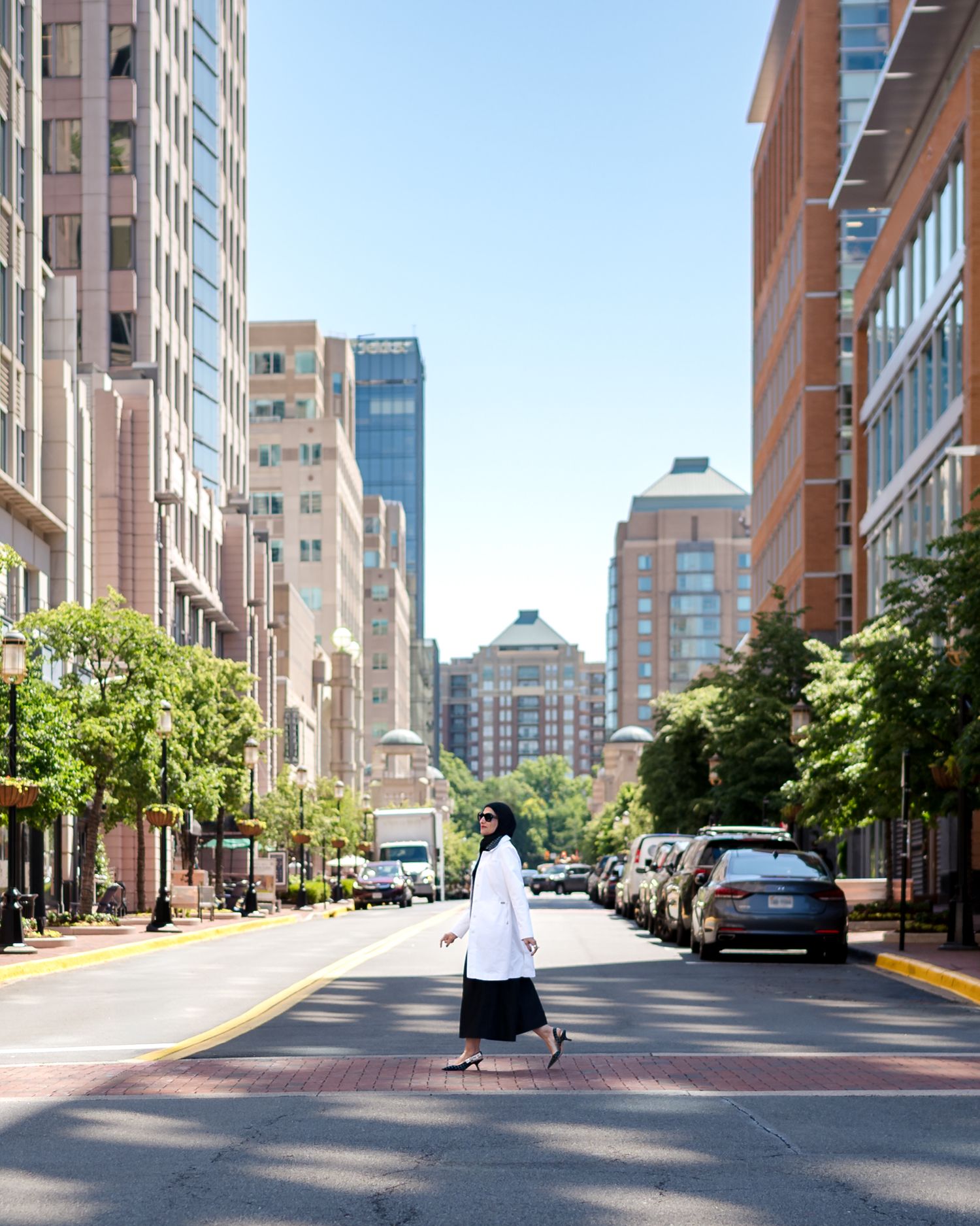 A woman in a white coat crosses a wide urban street lined with tall buildings and trees on a sunny day.