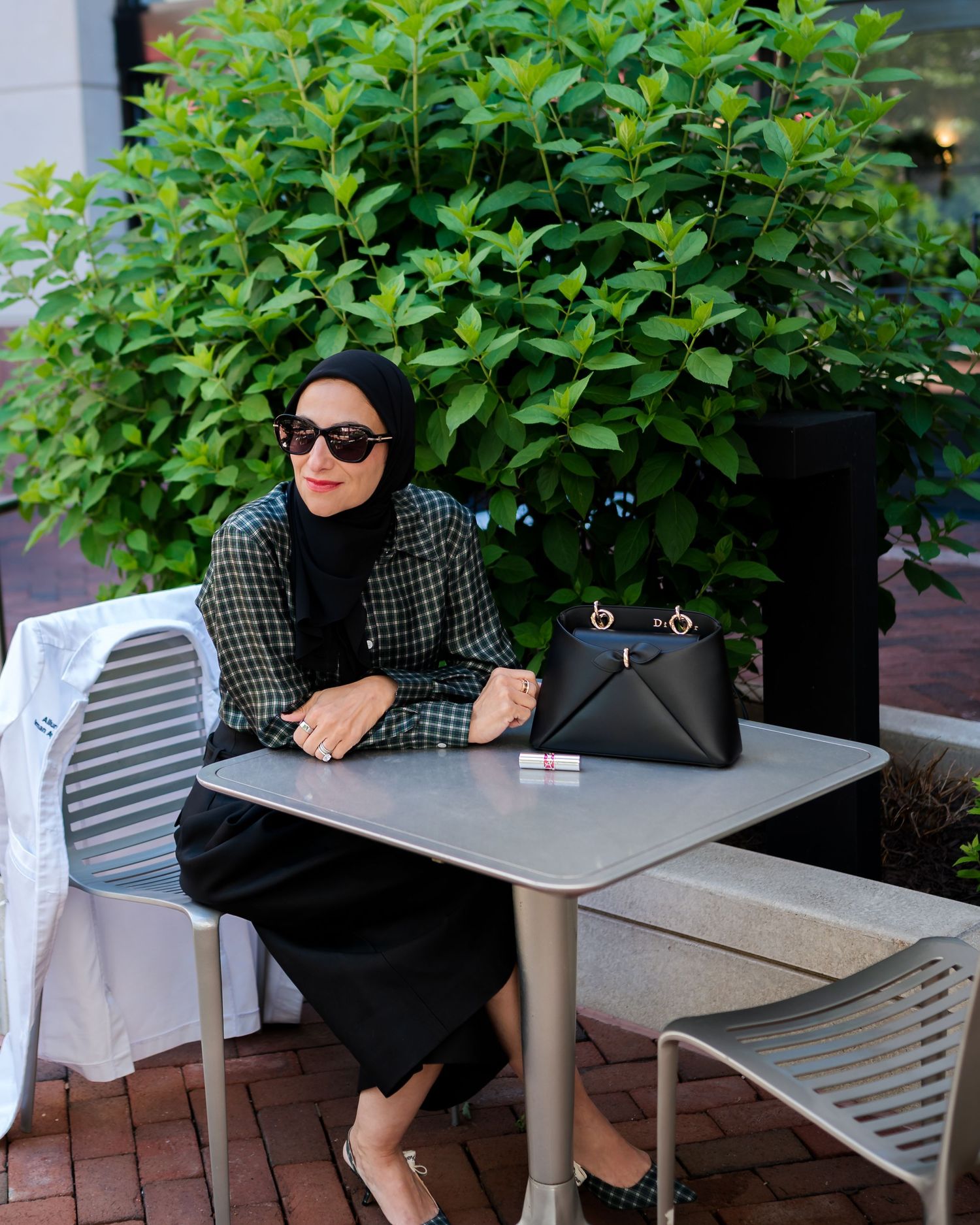 Woman in hijab wearing plaid blazer and black skirt sits at outdoor cafe table with designer handbag.