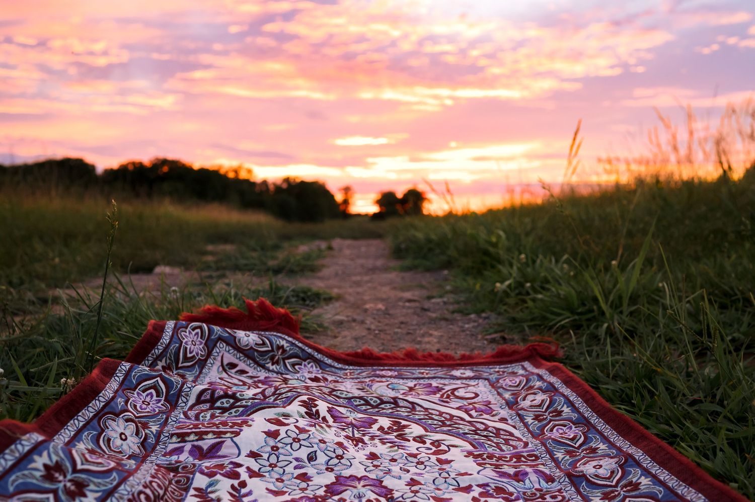A patterned blanket lies on a dirt path during a vibrant sunset with tall grass and trees in the background.
