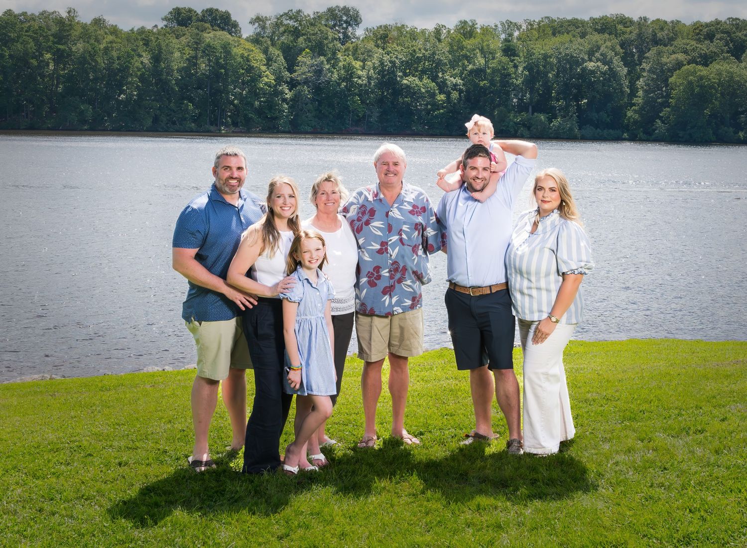 Large group poses together for outdoor family photos by serene lake during summer gathering.