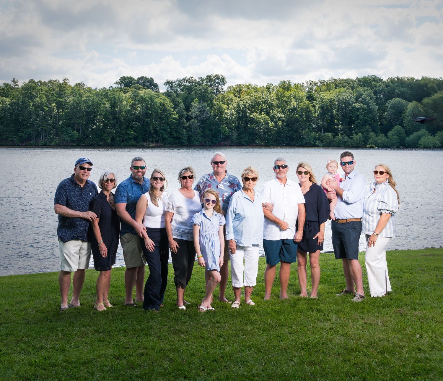 Wedding party in casual attire poses in a line along a scenic lakefront with trees in the background.