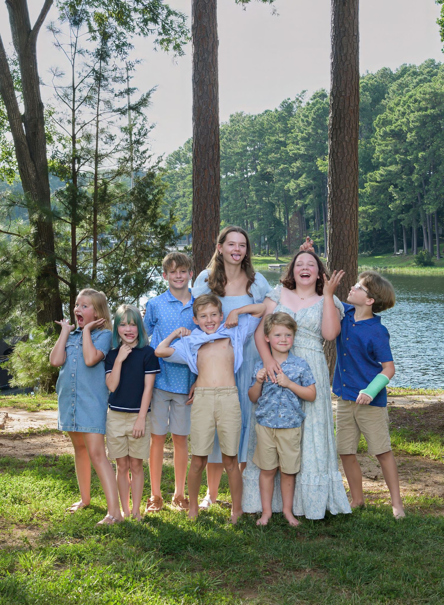 A group stands together laughing by a lakeside in summer, surrounded by tall pine trees and natural scenery.