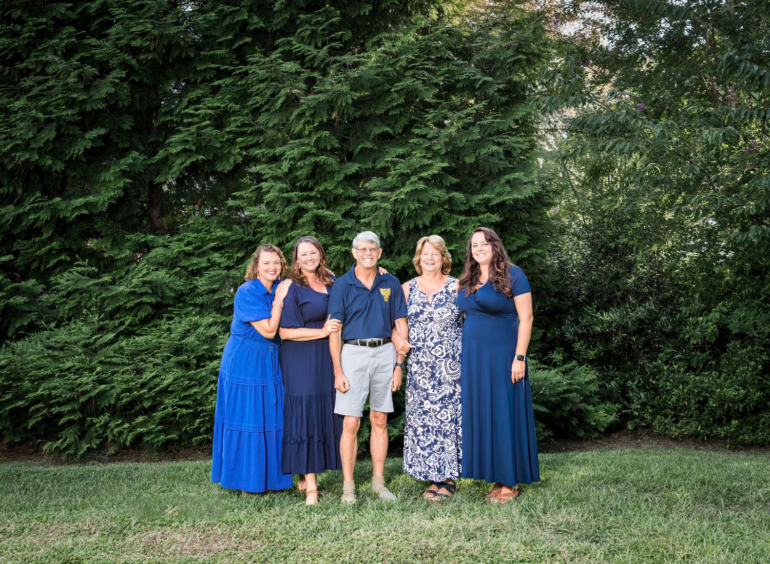 A group poses outdoors in coordinating navy blue and floral outfits against lush green foliage.