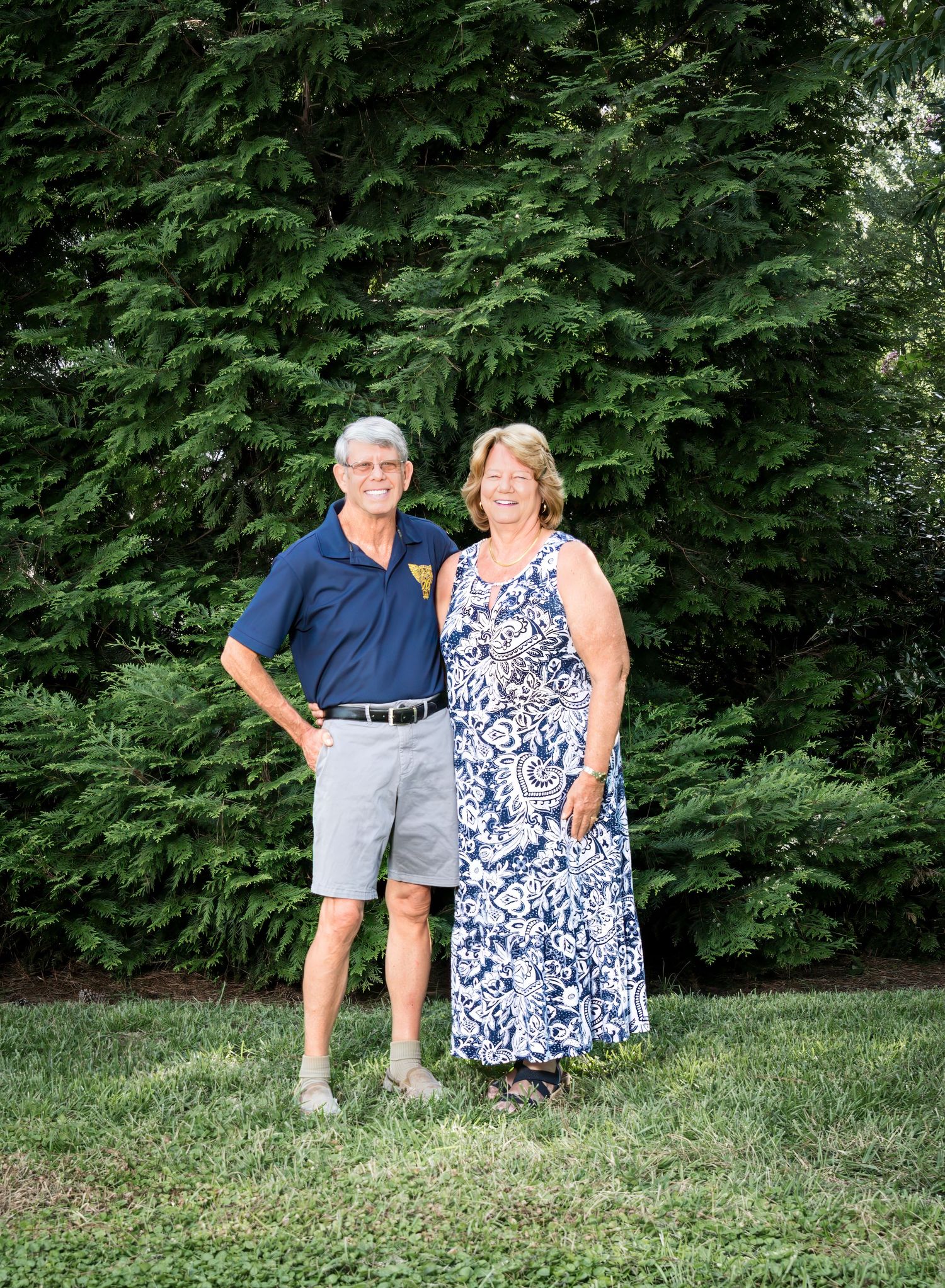 Two people pose together outdoors in front of lush green cedar trees on a summer day.