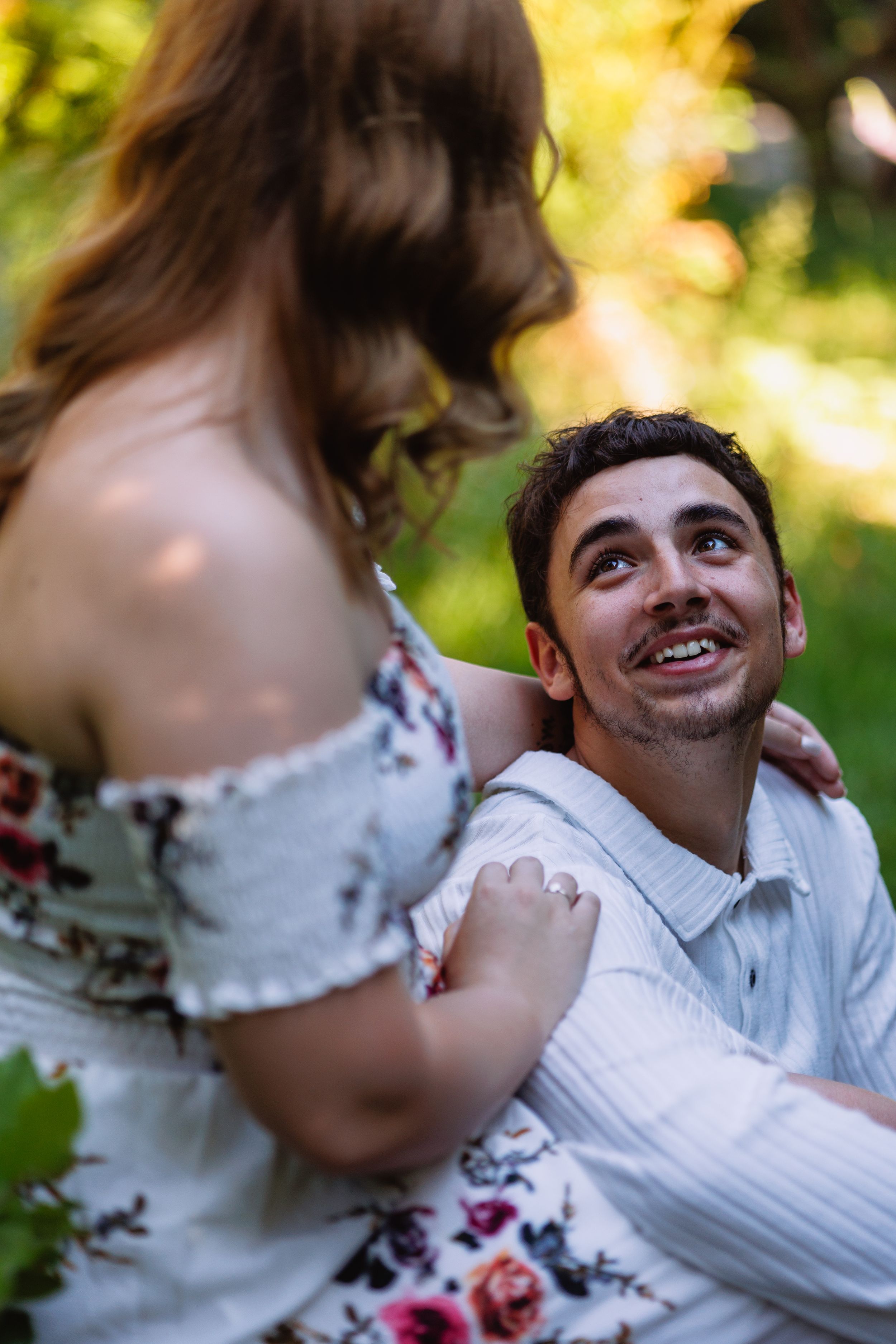 Smiling man gazes up adoringly at his partner during a sunny outdoor engagement photo session.