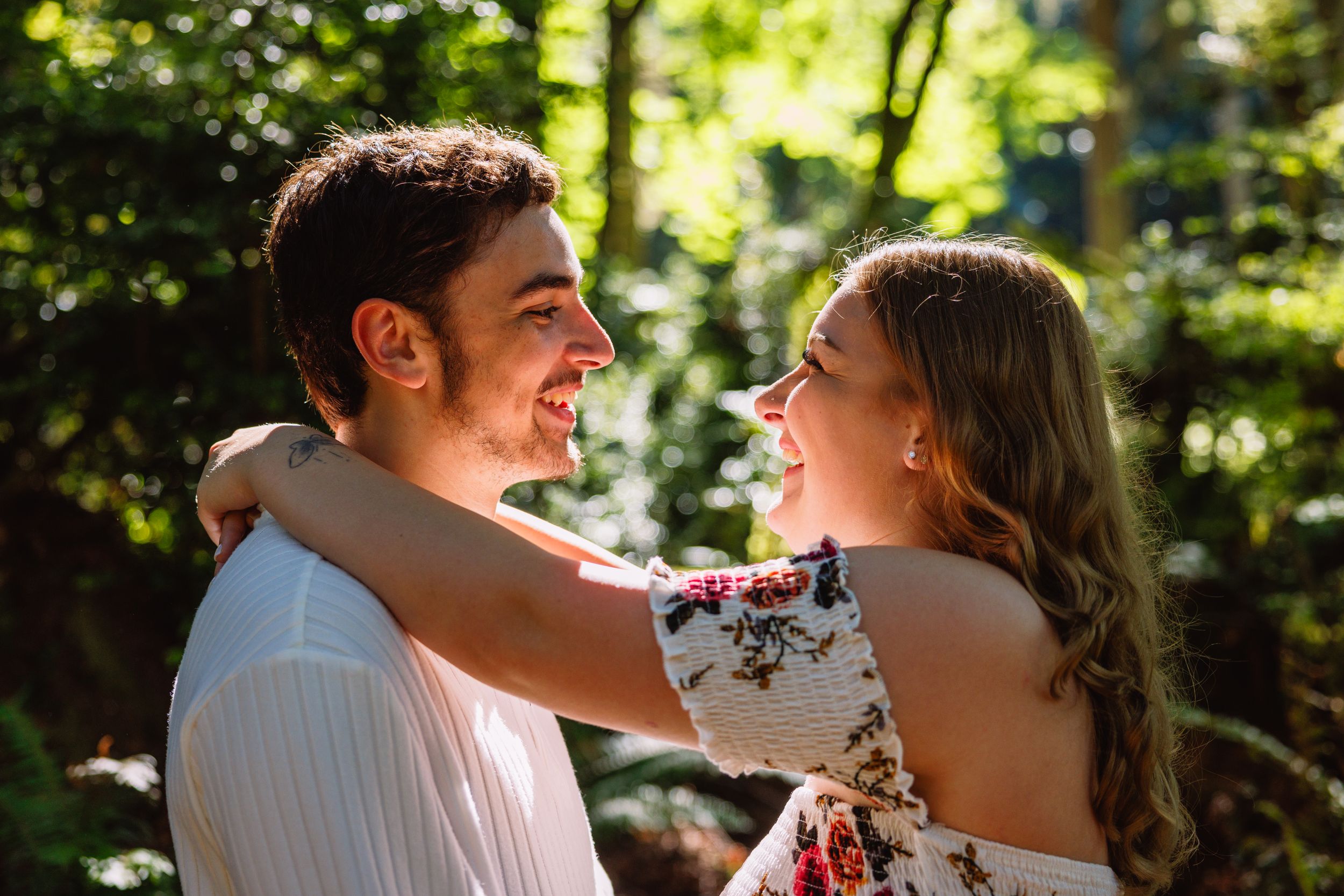 Happy couple embracing and laughing together in a sunlit forest during an engagement photoshoot.