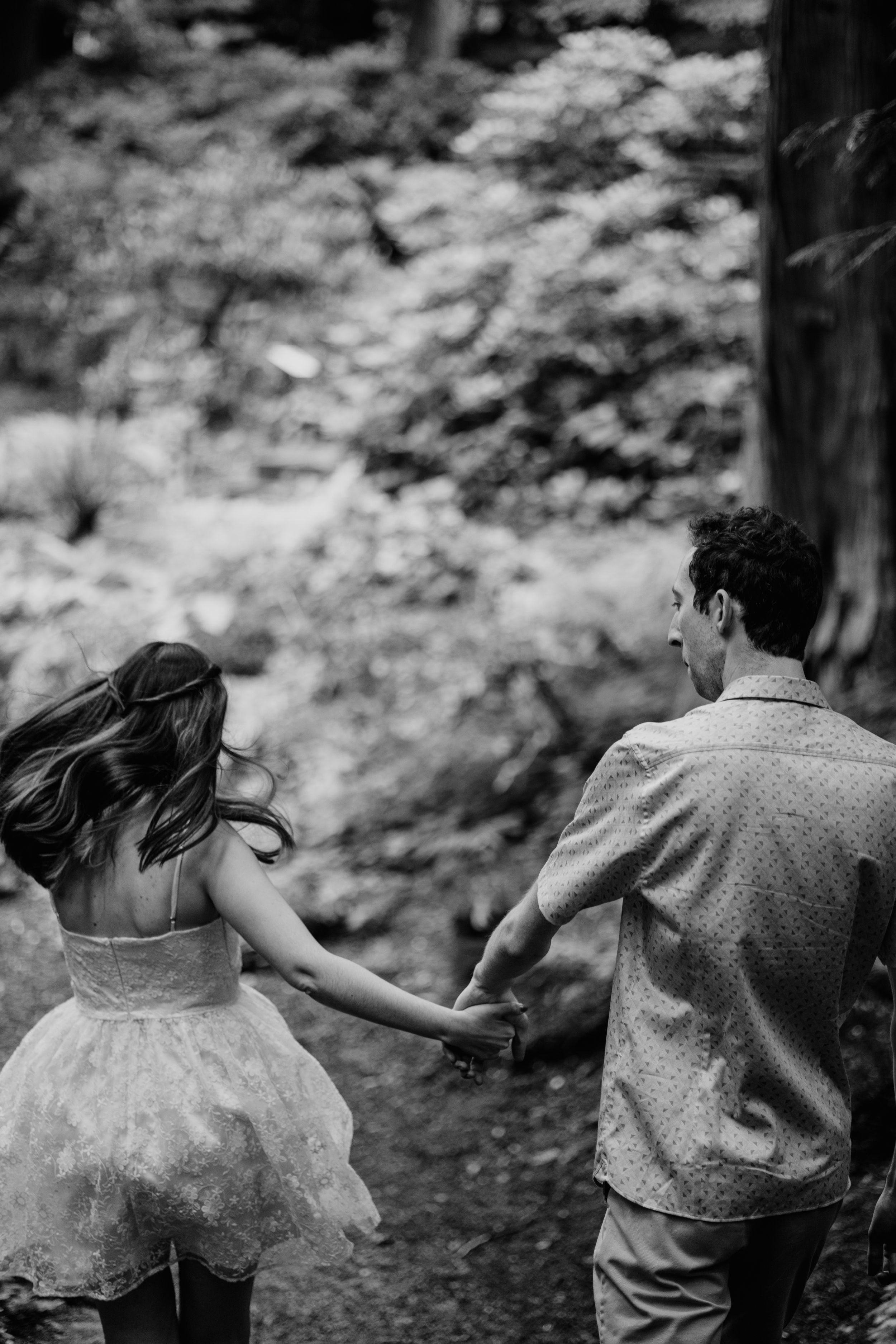 Black and white photo of couple walking hand in hand through a lush forest trail.