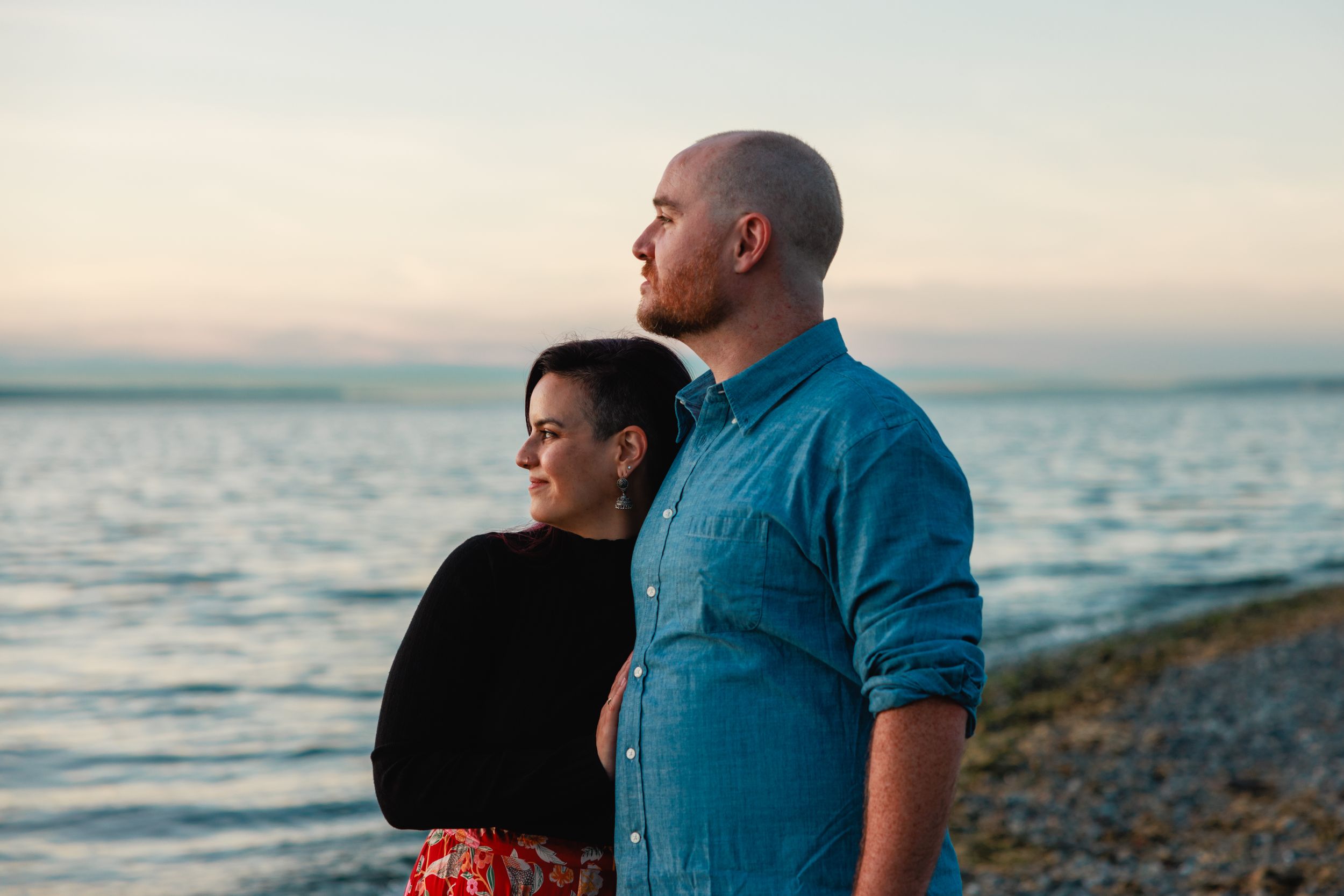 Couple standing together on a rocky beach at sunset, woman leaning against man in blue shirt.