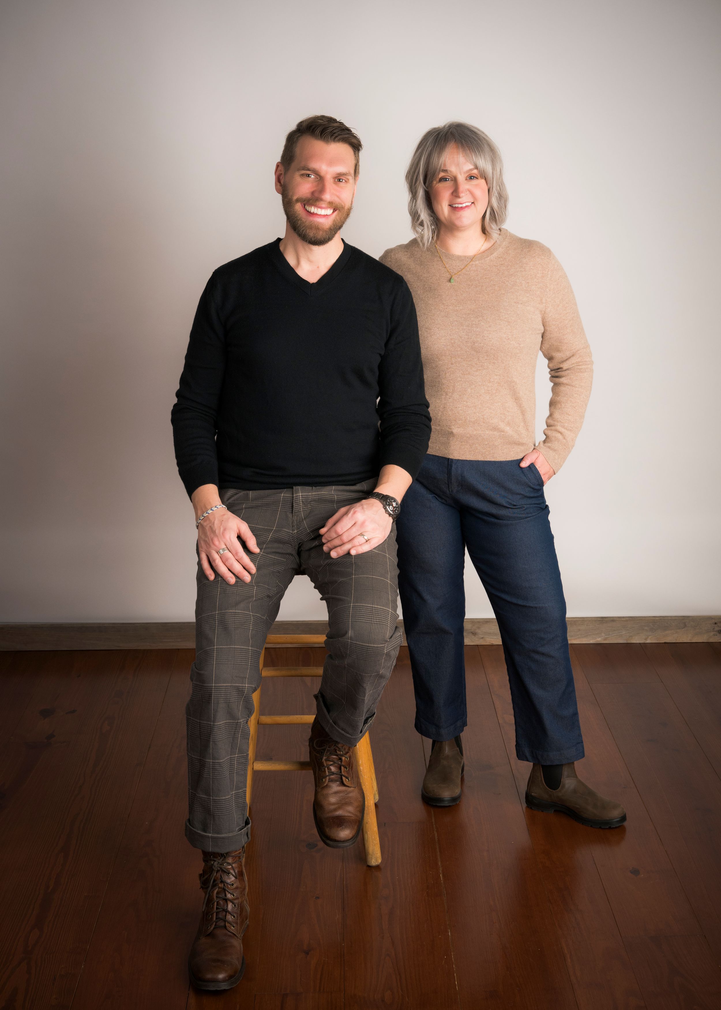 A smiling man and woman pose together in a bright studio, wearing casual sweaters and trousers for a professional portrait.
