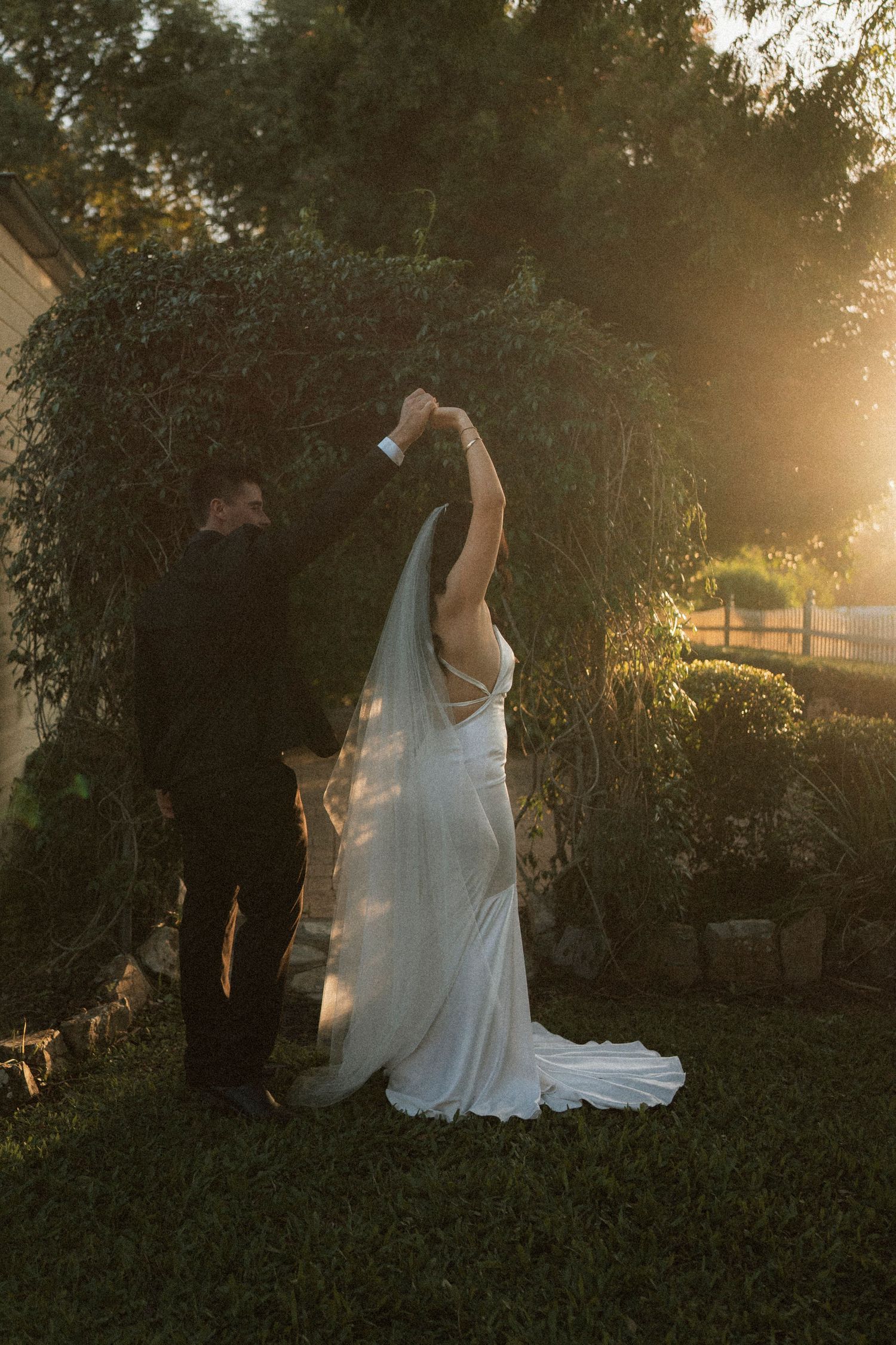 Bride and groom dancing at golden hour in a lush garden, her white gown and veil glowing in warm sunset light.