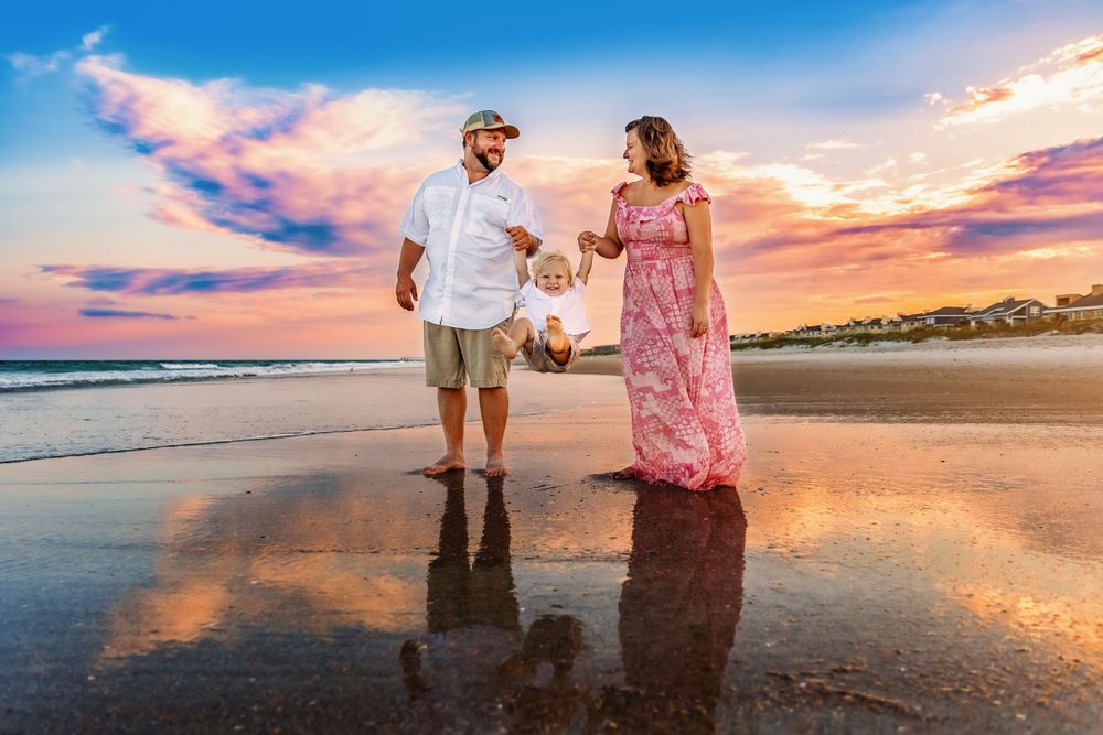 Two people walk hand in hand along a beach shoreline at sunset with colorful clouds reflecting in the wet sand.