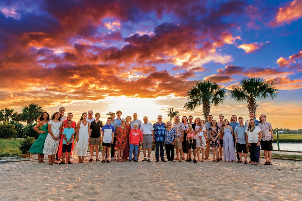 Large group poses for photo on beach with stunning orange and purple sunset sky.